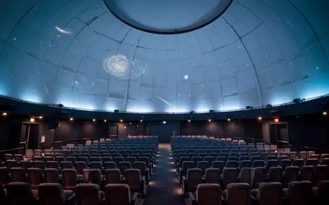 Innenansicht eines modernen Planetariums mit einer großen Kuppeldecke und Reihen von Sitzplätzen in gedimmtem Licht. - Foto: über boerse-global.de