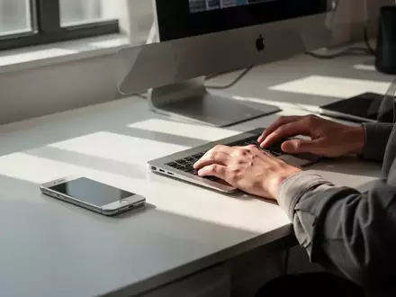 Person arbeitet an einem modernen Schreibtisch mit iPhone und MacBook in einer hellen, minimalistischen Umgebung. - Foto: über boerse-global.de