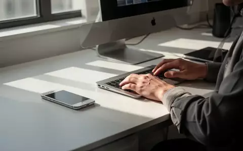 Person arbeitet an einem modernen Schreibtisch mit iPhone und MacBook in einer hellen, minimalistischen Umgebung. - Foto: über boerse-global.de