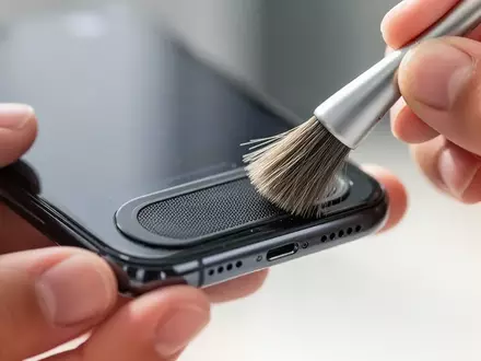 Nahaufnahme einer Person, die vorsichtig die Lautsprecheröffnungen eines iPhones mit einer weichen Bürste reinigt. - Foto: über boerse-global.de