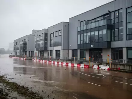 Moderne Industriegebäude mit Hochwasserschutzbarrieren an einem Flussufer bei bewölktem Himmel. - Foto: über boerse-global.de