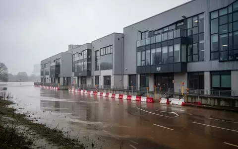 Moderne Industriegebäude mit Hochwasserschutzbarrieren an einem Flussufer bei bewölktem Himmel. - Foto: über boerse-global.de