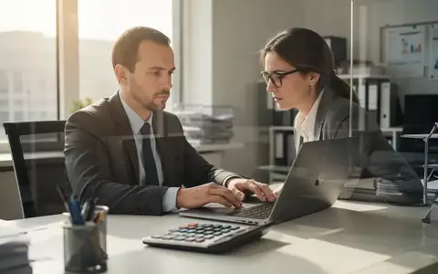 Eine Person arbeitet an einem Schreibtisch mit Taschenrechner und Laptop in einem hellen Büro. - Foto: über boerse-global.de
