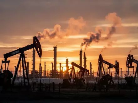 Ölförderanlagen bei Sonnenuntergang vor einer industriellen Raffinerie. - Foto: über boerse-global.de