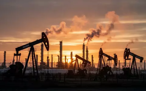 Ölförderanlagen bei Sonnenuntergang vor einer industriellen Raffinerie. - Foto: über boerse-global.de