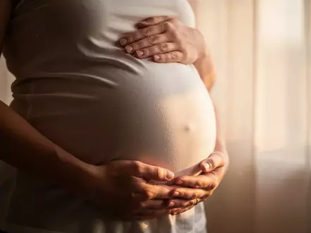 Nahaufnahme der Hände einer schwangeren Frau, die sanft ihren Bauch halten, in weichem Tageslicht. - Foto: über boerse-global.de