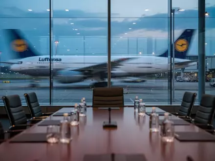 Ein Konferenztisch vor einem Fenster mit Blick auf ein Lufthansa-Flugzeug auf dem Rollfeld bei Dämmerung. - Foto: über boerse-global.de