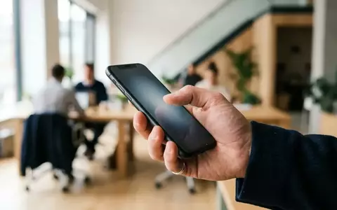Eine Hand hält ein modernes Smartphone, während ein Finger auf den Bildschirm tippt, in einer hellen Büroumgebung. - Foto: über boerse-global.de