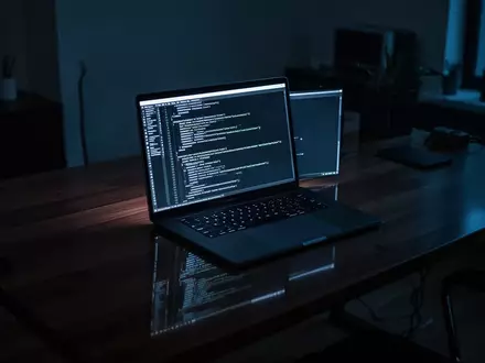 Ein minimalistischer Arbeitsplatz mit einem Laptop, auf dem abstrakte Programmiercodes in einem dunklen Raum zu sehen sind. - Foto: über boerse-global.de