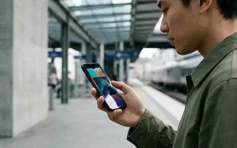 Eine Person hält ein Smartphone in einem Bahnhof, auf dessen Display ein farbenfrohes, digitales Ticket angezeigt wird. - Foto: über boerse-global.de