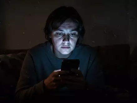 Eine einsame Person blickt in einem abgedunkelten Raum auf ein leuchtendes Smartphone, das ihr Gesicht beleuchtet. - Foto: über boerse-global.de