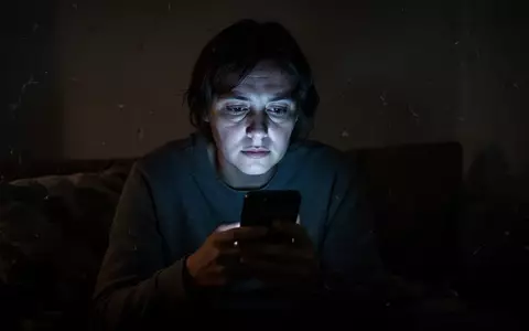 Eine einsame Person blickt in einem abgedunkelten Raum auf ein leuchtendes Smartphone, das ihr Gesicht beleuchtet. - Foto: über boerse-global.de