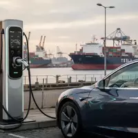 Moderne Ladesäule für Elektroautos vor einer industriellen Hafenanlage mit Containerschiffen im Hintergrund. - Foto: über boerse-global.de