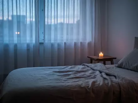 Minimalistisches Schlafzimmer in gedimmtem Abendlicht mit einem unangezündeten Kerzenlicht auf einem Nachttisch. - Foto: über boerse-global.de