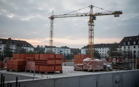 Eine verlassene Baustelle mit einem unbesetzten Baukran und gestapelten Baumaterialien vor einem bewölkten Himmel. - Foto: über boerse-global.de