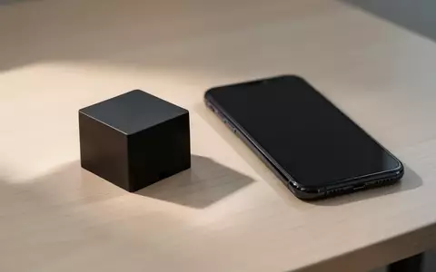Ein kleiner, schwarzer Plastikblock liegt neben einem modernen Smartphone mit ausgeschaltetem Bildschirm auf einem Holztisch. - Foto: über boerse-global.de