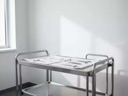 Ein minimalistisch eingerichteter, heller Behandlungsraum in einer Arztpraxis mit einem leeren medizinischen Gerätewagen. - Foto: über boerse-global.de