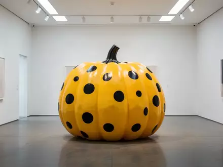 Eine große, gelbe Kürbisskulptur mit schwarzen Punkten steht in einem hellen, minimalistischen Kunstraum. - Foto: über boerse-global.de