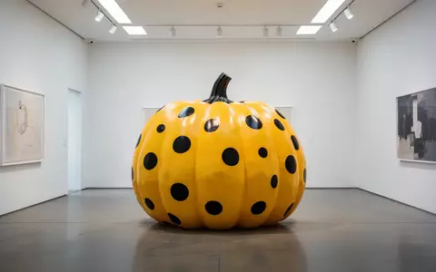 Eine große, gelbe Kürbisskulptur mit schwarzen Punkten steht in einem hellen, minimalistischen Kunstraum. - Foto: über boerse-global.de
