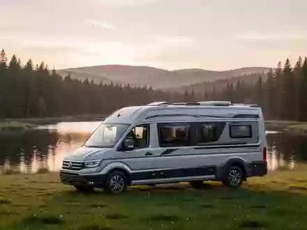 Moderner Campingbus geparkt auf einer Wiese vor einem Seeufer in einer hügeligen Landschaft bei Sonnenaufgang. - Foto: über boerse-global.de