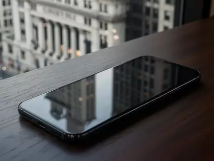 Ein unbenutztes Smartphone auf einem dunklen Schreibtisch vor einer verschwommenen Bankfiliale. - Foto: über boerse-global.de