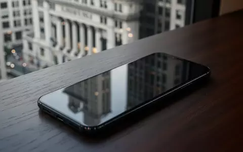 Ein unbenutztes Smartphone auf einem dunklen Schreibtisch vor einer verschwommenen Bankfiliale. - Foto: über boerse-global.de