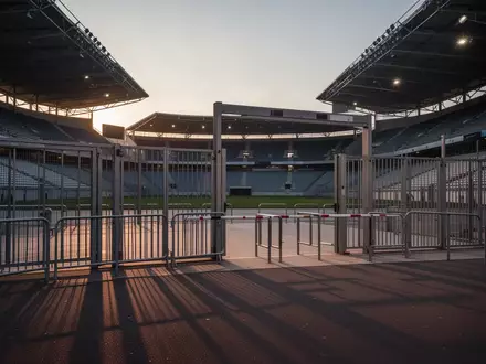 Leerer Eingangsbereich eines modernen Stadions mit Sicherheitsbarrieren und Metallzäunen bei Dämmerung. - Foto: über boerse-global.de