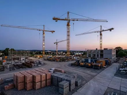 Inaktive Baukräne auf einer Baustelle vor einem klaren Abendhimmel, davor gestapelte Baumaterialien. - Foto: über boerse-global.de