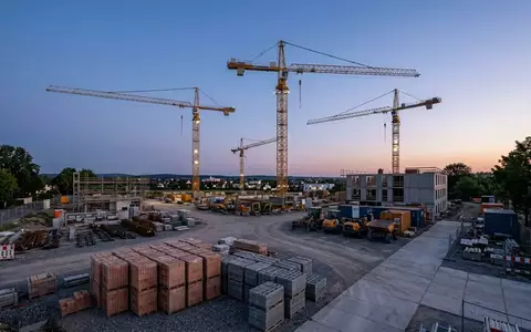 Inaktive Baukräne auf einer Baustelle vor einem klaren Abendhimmel, davor gestapelte Baumaterialien. - Foto: über boerse-global.de