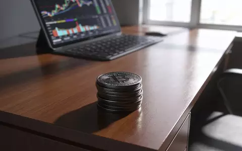 Metallische Kryptowährungstoken auf einem Schreibtisch vor einem unscharfen Finanzterminal. - Foto: über boerse-global.de