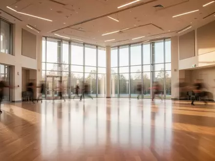 Ein lichtdurchfluteter, leerer Tanzsaal mit poliertem Parkettboden und großen Fensterfronten in einer modernen Einrichtung. - Foto: über boerse-global.de