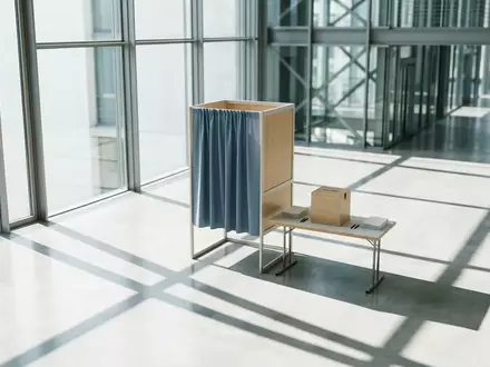 Eine leere Wahlkabine in einem modernen, lichtdurchfluteten Regierungsgebäude mit geometrischen Schattenwürfen. - Foto: über boerse-global.de