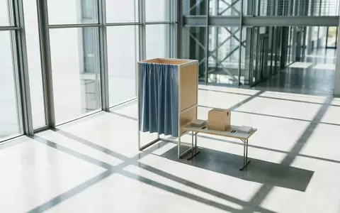 Eine leere Wahlkabine in einem modernen, lichtdurchfluteten Regierungsgebäude mit geometrischen Schattenwürfen. - Foto: über boerse-global.de