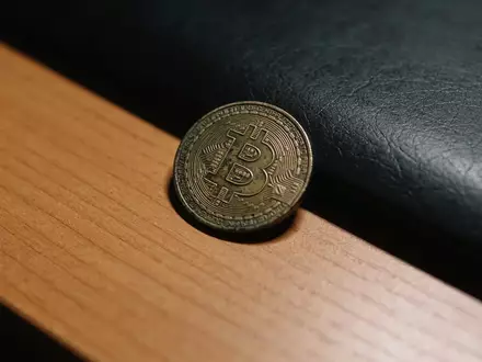 Ein einzelner Bitcoin-Token liegt einsam auf einem dunklen Schreibtisch in einem schummrig beleuchteten Raum. - Foto: über boerse-global.de