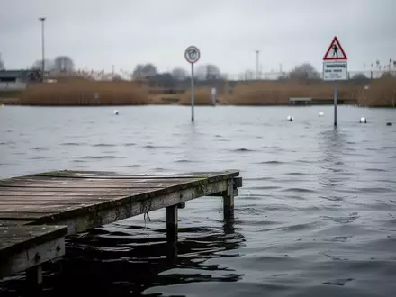 Ein ruhiger Badesee mit einem hölzernen Steg und bewölktem Himmel bei einer abgesperrten Wasserstelle. - Foto: über boerse-global.de