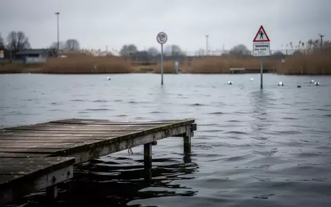 Ein ruhiger Badesee mit einem hölzernen Steg und bewölktem Himmel bei einer abgesperrten Wasserstelle. - Foto: über boerse-global.de