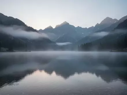 Eine neblige Berglandschaft bei Sonnenaufgang über einem spiegelglatten Bergsee, die Ruhe und Stille ausstrahlt. - Foto: über boerse-global.de