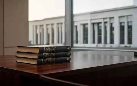 Stapel juristischer Fachbücher auf einem Schreibtisch vor einem unscharfen Hintergrund moderner Gerichtsarchitektur. - Foto: über boerse-global.de