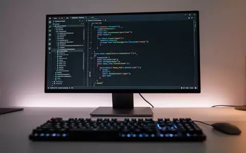 Ein moderner, minimalistischer Arbeitsplatz mit einem Monitor, auf dem eine dunkle Code-Umgebung zu sehen ist. - Foto: über boerse-global.de