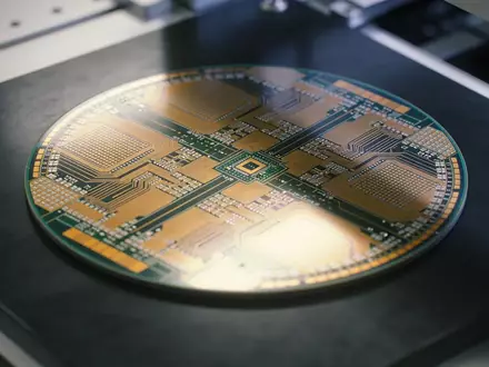 Ein Silizium-Wafer mit Mikrochip-Strukturen liegt auf einer dunklen Werkbank in einer Fertigungshalle. - Foto: über boerse-global.de