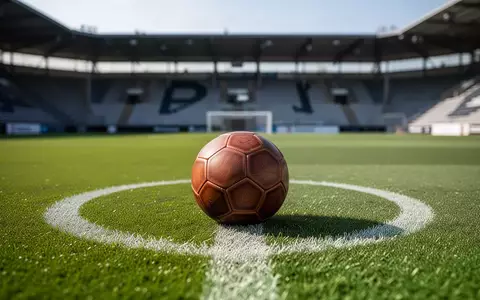 Ein klassischer Fußball liegt auf dem grünen Rasen eines Sportplatzes vor leeren Zuschauerrängen. - Foto: über boerse-global.de