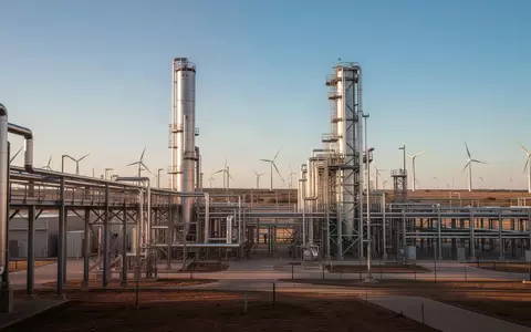 Moderne Industrieanlage in Texas bei Sonnenuntergang mit Metallinfrastruktur und Windkraftanlagen. - Foto: über boerse-global.de