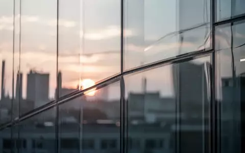 Moderne, minimalistische Glasfassade mit fließenden, transparenten Reflexionen bei Sonnenuntergang. - Foto: über boerse-global.de