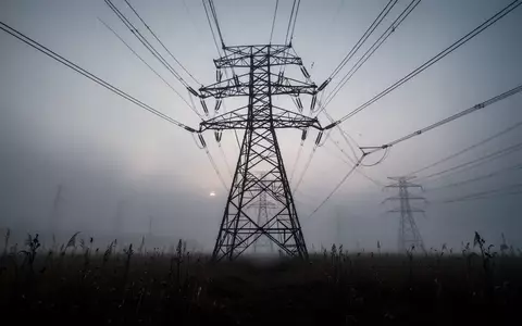 Ein massiver Hochspannungsmast auf einem nebligen Feld in der Dämmerung, der die kritische Energieinfrastruktur symbolisiert. - Foto: über boerse-global.de