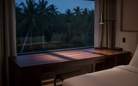 Ein minimalistisch eingerichtetes Hotelzimmer bei Dämmerung mit einem leeren Schreibtisch und Blick auf eine tropische Landschaft. - Foto: über boerse-global.de