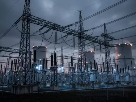 Ein weites, atmosphärisches Foto eines modernen Umspannwerks in der Dämmerung mit komplexen Hochspannungsleitungen und Industriestrukturen unter einem bewölkten Himmel. - Foto: über boerse-global.de