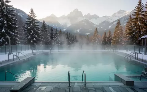 Ein ruhiges, leeres Thermalbecken im Freien vor einer alpinen Bergkulisse mit aufsteigendem Dampf im Morgenlicht. - Foto: über boerse-global.de