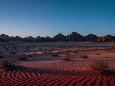 Weite, karge Wüstenlandschaft bei Dämmerung mit tiefblauem Himmel und schroffen Felsformationen in der Ferne. - Foto: über boerse-global.de