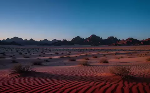Weite, karge Wüstenlandschaft bei Dämmerung mit tiefblauem Himmel und schroffen Felsformationen in der Ferne. - Foto: über boerse-global.de