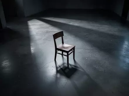 Ein leerer Holzstuhl in einem minimalistischen Raum mit langen Schatten auf einem Betonboden, der die Atmosphäre von finanzieller Verletzlichkeit unterstreicht. - Foto: über boerse-global.de